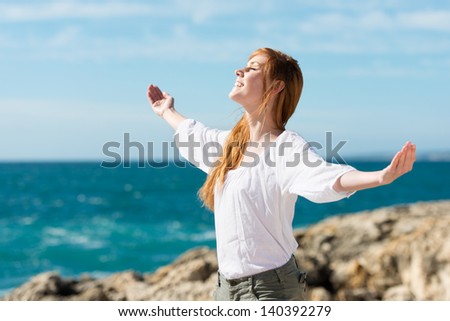 A pretty happy young woman enjoying the sun stands with her arms outspread against an ocean backdrop
