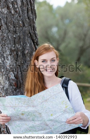 Woman finding her route on a map leaning up against the trunk of a tree with a rucksack on her back