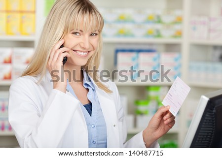 Portrait of happy female pharmacist holding prescription paper while using mobilephone in pharmacy