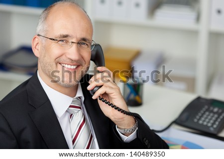 Portrait of happy businessman using landline phone at office desk
