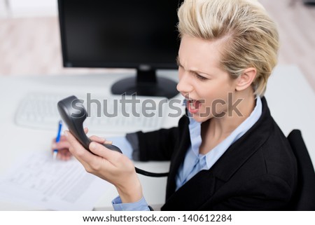 Frustrated young businesswoman shouting on telephone receiver at office desk