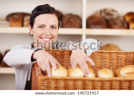 Smiling attractive female assistant selecting rolls in a bakery from a large wicker basket and smiling at the camera