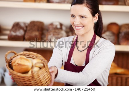 Woman Working In A Bakery Offering A Customer A Basket Of Assorted Bread And Rolls With A Friendly Smile, Focus To The Woman