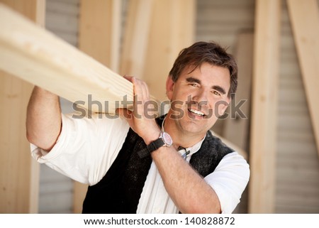 Smiling mature carpenter carrying a large wood plank on his shoulder