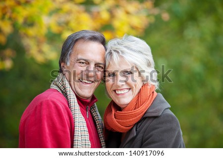 Head and shoulders portrait of a joyful attractive senior couple standing in front of a colourful yellow autumn tree dressed in trendy scarves