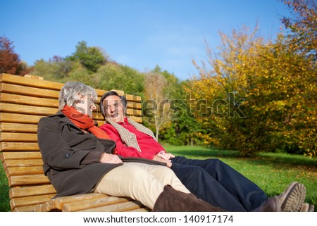 Senior couple relaxing in the autumn sun reclining side by side on a comfortable curved wooden bench in a park or garden