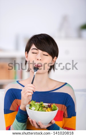 Woman enjoying a bowl of fresh leafy green salad salad standing with her eyes closed in pleasure and a fork in her mouth