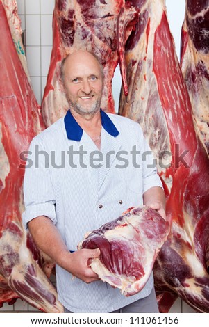 Happy middle-aged butcher holding a freshly cut piece of beef near a hanging cow carcass inside a butchery