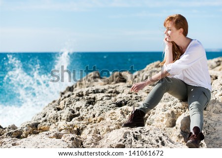 Thoughtful casual young woman relaxing at the sea sitting on the rocks watching the spray from the breaking waves