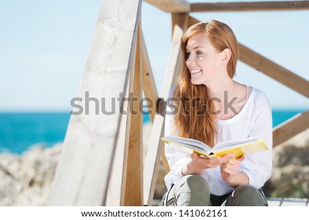 Beautiful young woman relaxing with a book at the sea sitting on a flight of wooden steps looking at the ocean