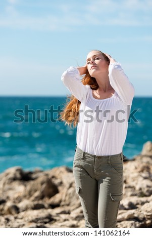 Pretty woman enjoying the sun on her face standing with her back to the ocean on a rocky shore holding back her hair off her face with her hands