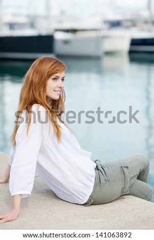 Pretty casual young woman sitting relaxing on a harbour wall with moored boats in the background