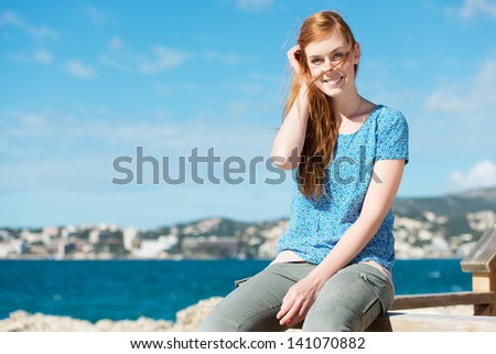 Young woman enjoying life at the sea sitting on a wooden rail against an ocean backdrop smiling at the camera