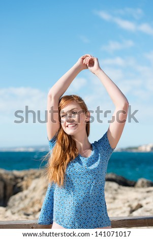 Carefree beautiful young woman at the seaside enjoying the warm summer sun standing with her arms raised above her head