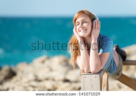 Pretty young woman wearing headphones lying on a wooden railing listening to music at the sea