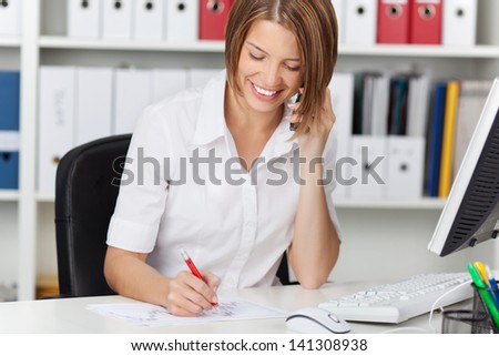 Beautiful young businesswoman calling by phone while writing on white paper at office