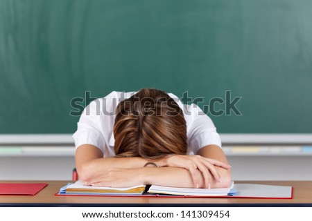 Teacher suffering from acute stress resting her head on her arms at her desk in front of the blackboard as she seeks to gather herself together