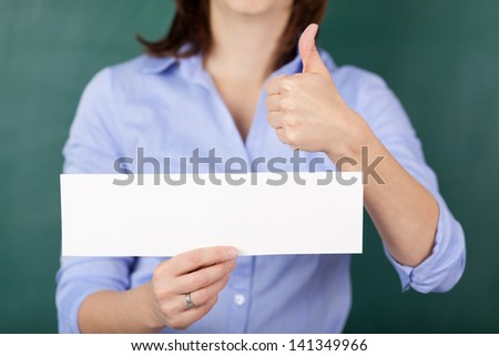 Midsection of female teacher holding blank paper while gesturing thumbs up against chalkboard
