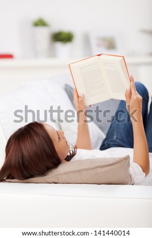 Woman lying reading on a couch with the top of her head facing the camera and the book in the air