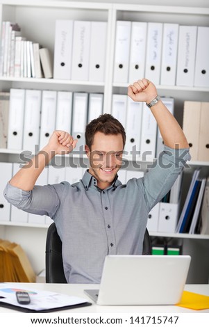 Happy young businessman raising hands in front of laptop at office desk