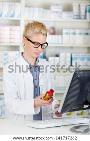 Young female pharmacist checking prescription bottle by computer at the drugstore