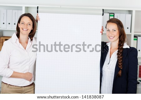 Two successful smiling businesswomen giving a flip chart presentation standing on either side of a blank white sheet of paper