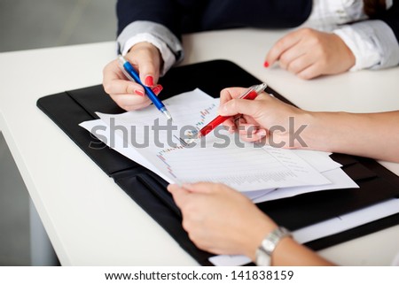 Cropped view of the hands of two women in a business meeting sitting at a table discussing a document and graph while making notes on it with their pens