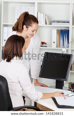 Two female office workers working together on a computer in the office with the blank screen visible to the viewer