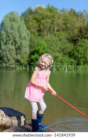 Pretty little girl fishing at the lake standing on the rocky edge in her gumboots dangling her net in the water