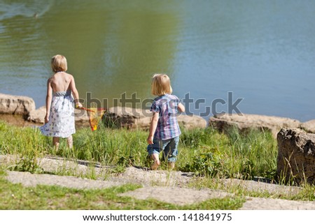 Two little girls with their backs to the camera fishing at the lake with a small net in summer sunshine