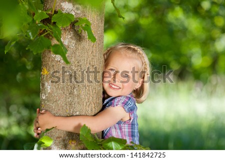 Happy little girl hugging a tree standing with her arms around the trunk smiling at the camera