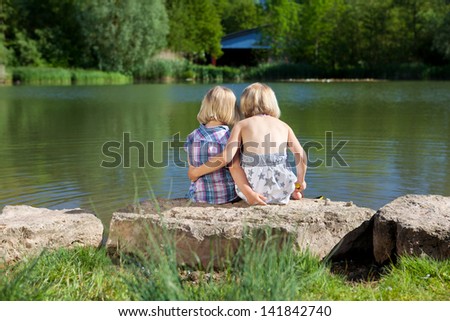 Two loving little sisters at the lake sitting with their arms around each other facing away from the camera on a rock on the shore