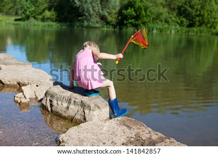 Little girl climbing into the lake off a rock in her gumboots with a small fishing net in her hand, rear view