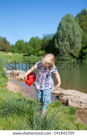 Little girl playing in a natural environment standing at the side of a tranquil lake watering the reeds with a plastic watering can
