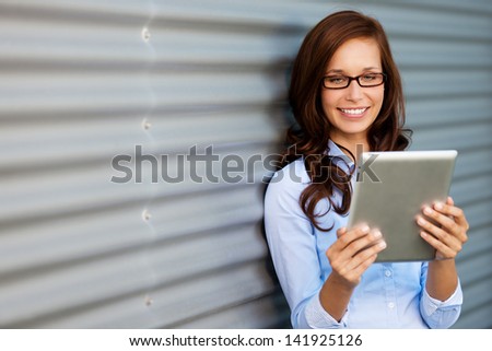 Young woman wearing glasses leaning against a corrugated metal wall or door reading her tablet computer, angled with copyspace