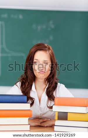 Student eyeing her books with a rueful expression and one eye closed as she sits at her desk flanked on either side by a pile of books