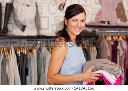 Portrait of happy smiling woman holding stack of clothes in boutique