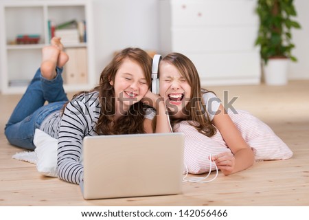Two Happy Young Teenage Girls Sharing Headphones Connected To A Laptop To Listen To Music While Laying On The Living-Room Floor