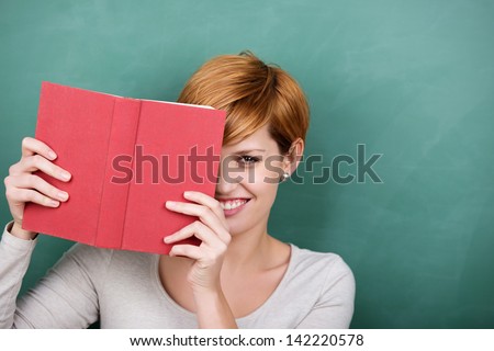 Portrait of female student peeking out of book against chalkboard