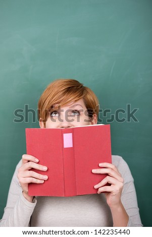 Portrait of female student peeking out of red book against chalkboard