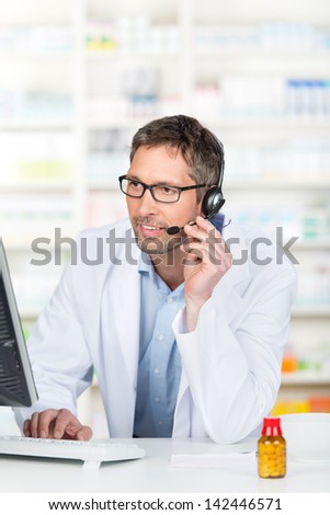 Mature male pharmacist wearing headset while using computer at pharmacy counter