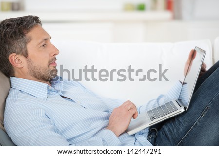 Side View Of Happy Mature Man Holding Laptop While Lying On Couch In House