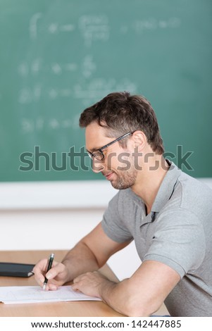 Side view of male teacher checking examination papers at bench in classroom