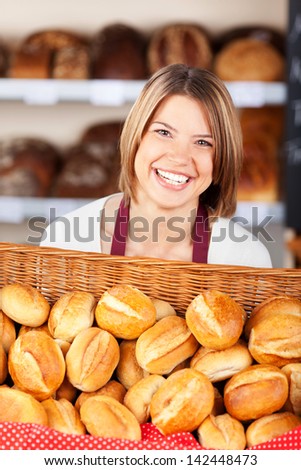 Smiling woman working in a bakery carrying a large flat wicker basket filled with crisp crusty golden rolls