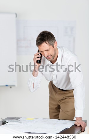 Mature male architect looking at blueprint while using cordless phone at desk in office