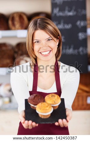 Female bakery worker with fresh muffins holding them out on a tray in front of her while smiling at the camera