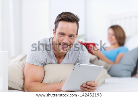 Smiling young man relaxing with a tablet-pc lying on his stomach on top of his bed having a lazy day