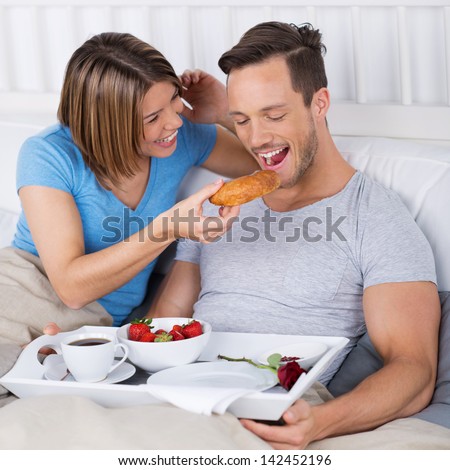 Laughing young couple enjoying breakfast in bed as the wife tries to feed her husband a croissant