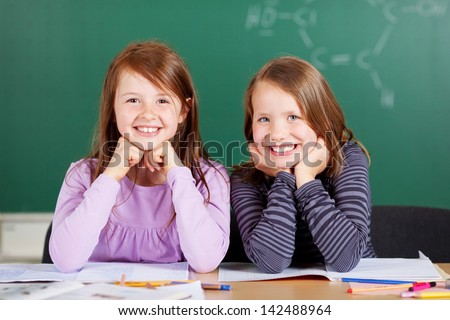 Two pretty smiling girls in school sitting close together behind the table with a chalkboard backdrop