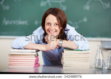 Smiling female teacher in class giving chemistry lessons ans leaning on two piles of textbooks looking at the camera